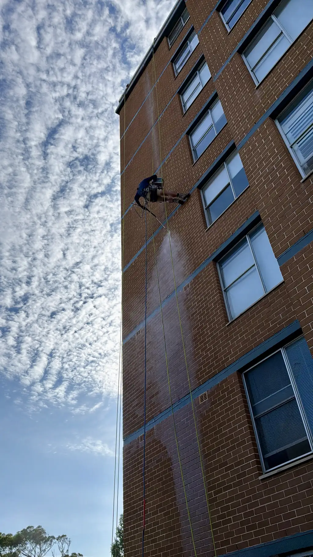 Rope access technician pressure-washing a brick facade