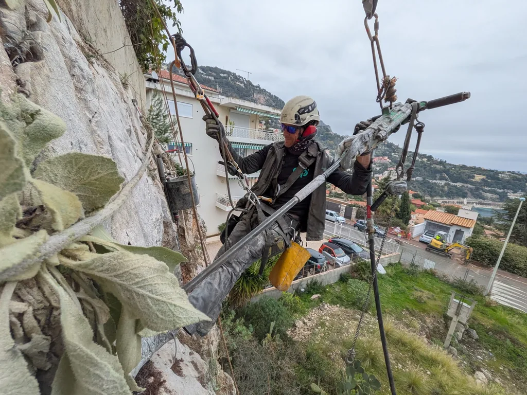 Rope access technician with a rotary hammer on a rock face above the town