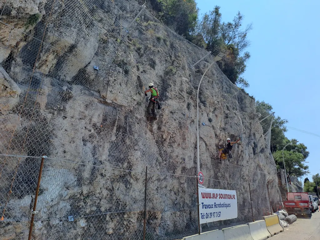 Rope access technicians on a meshed rock face beside a road