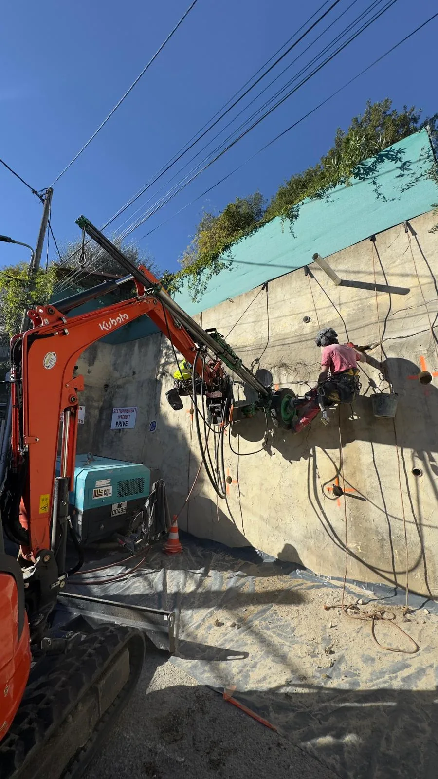 Rope access technician and mini excavator drilling on a retaining wall